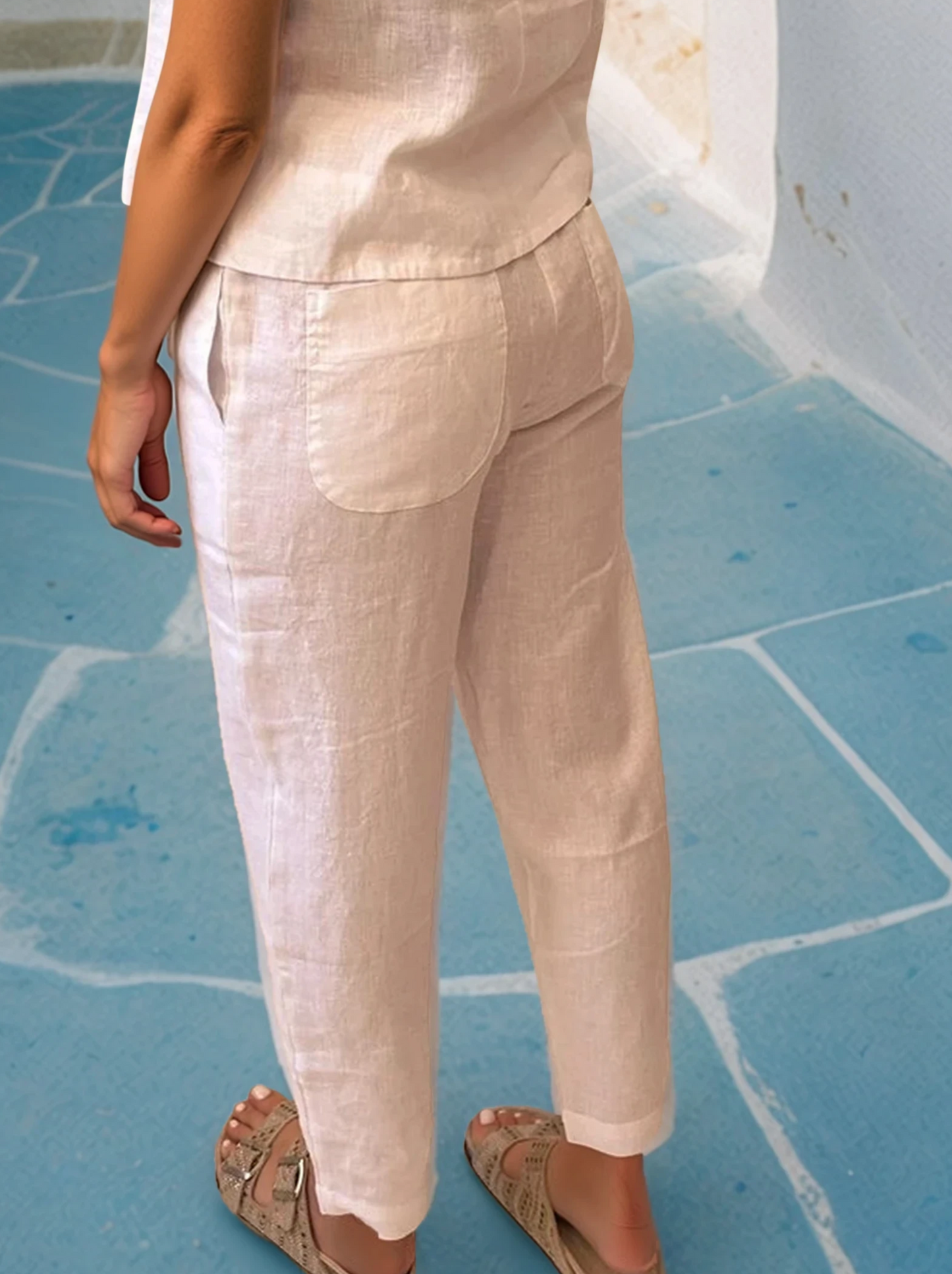 Model wearing relaxed-fit tapered linen trousers with side and back pockets, crafted in Greece from 100% linen, styled in a whitewashed Cycladic alley.