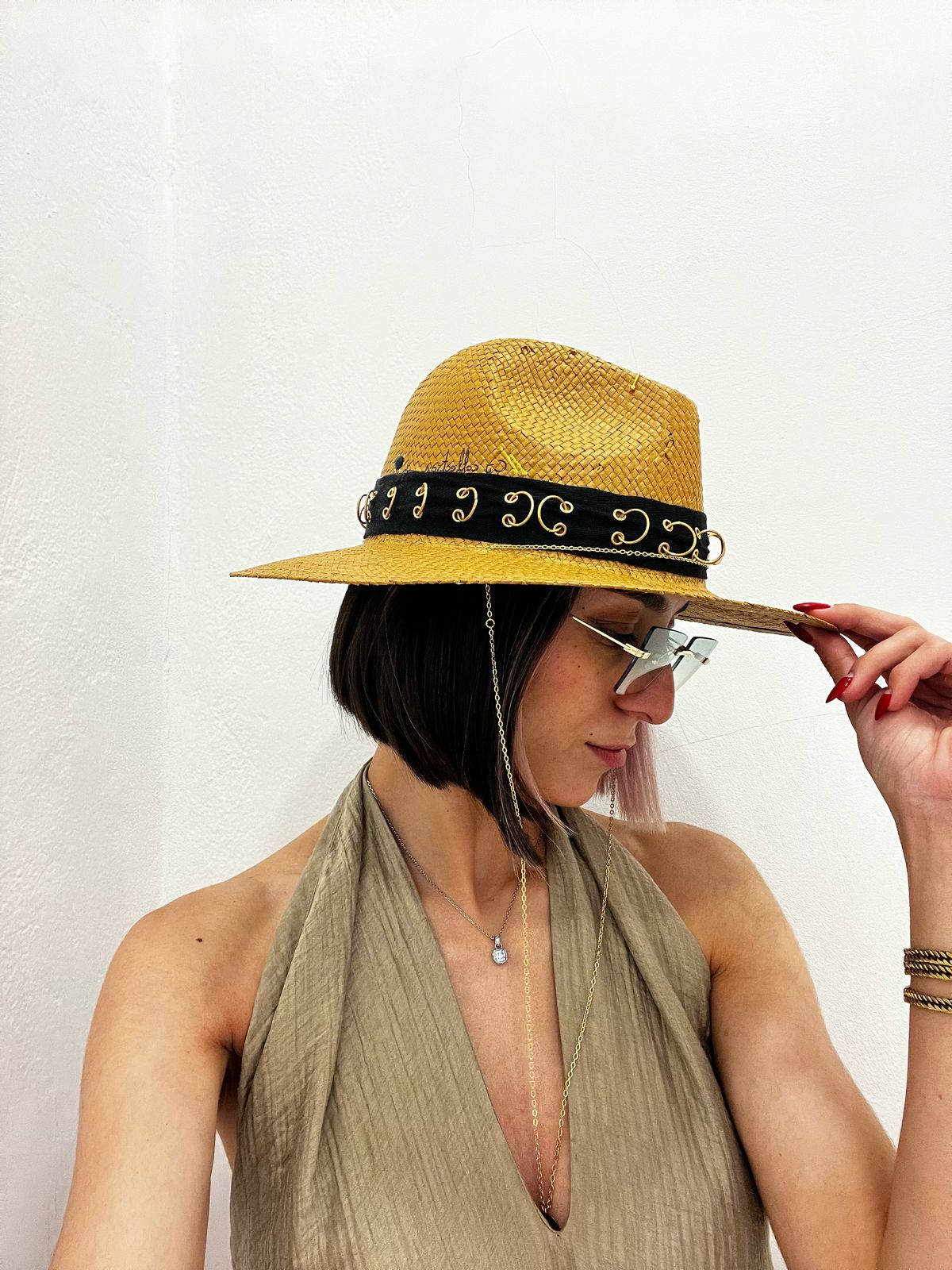 Woman wearing a woven straw hat with a black and gold embellished band, styled with sunglasses and a halter top, photographed in a minimalist indoor setting.