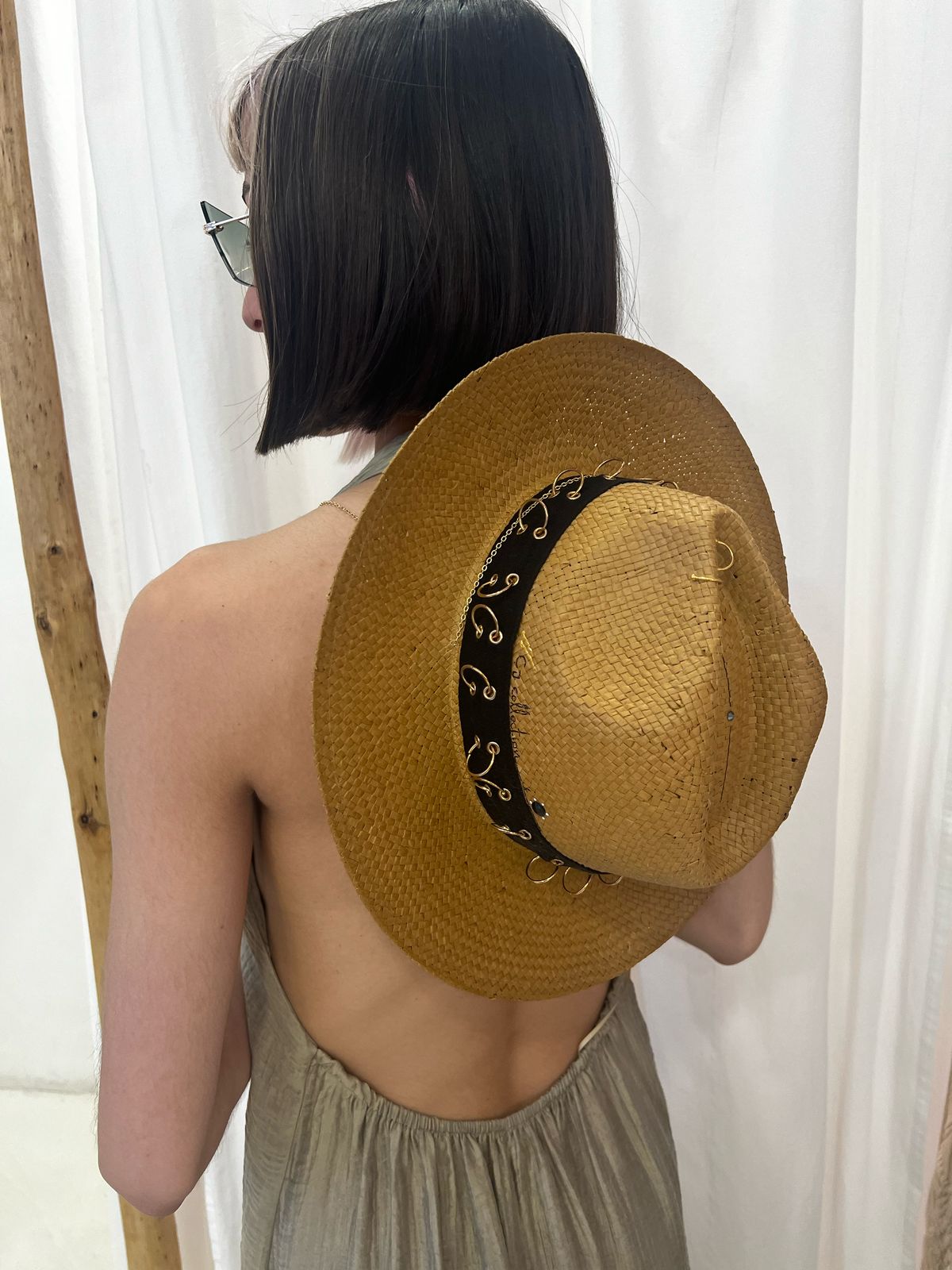 Woman wearing a woven straw hat with a black and gold embellished band, styled with sunglasses and a halter top, photographed in a minimalist indoor setting.