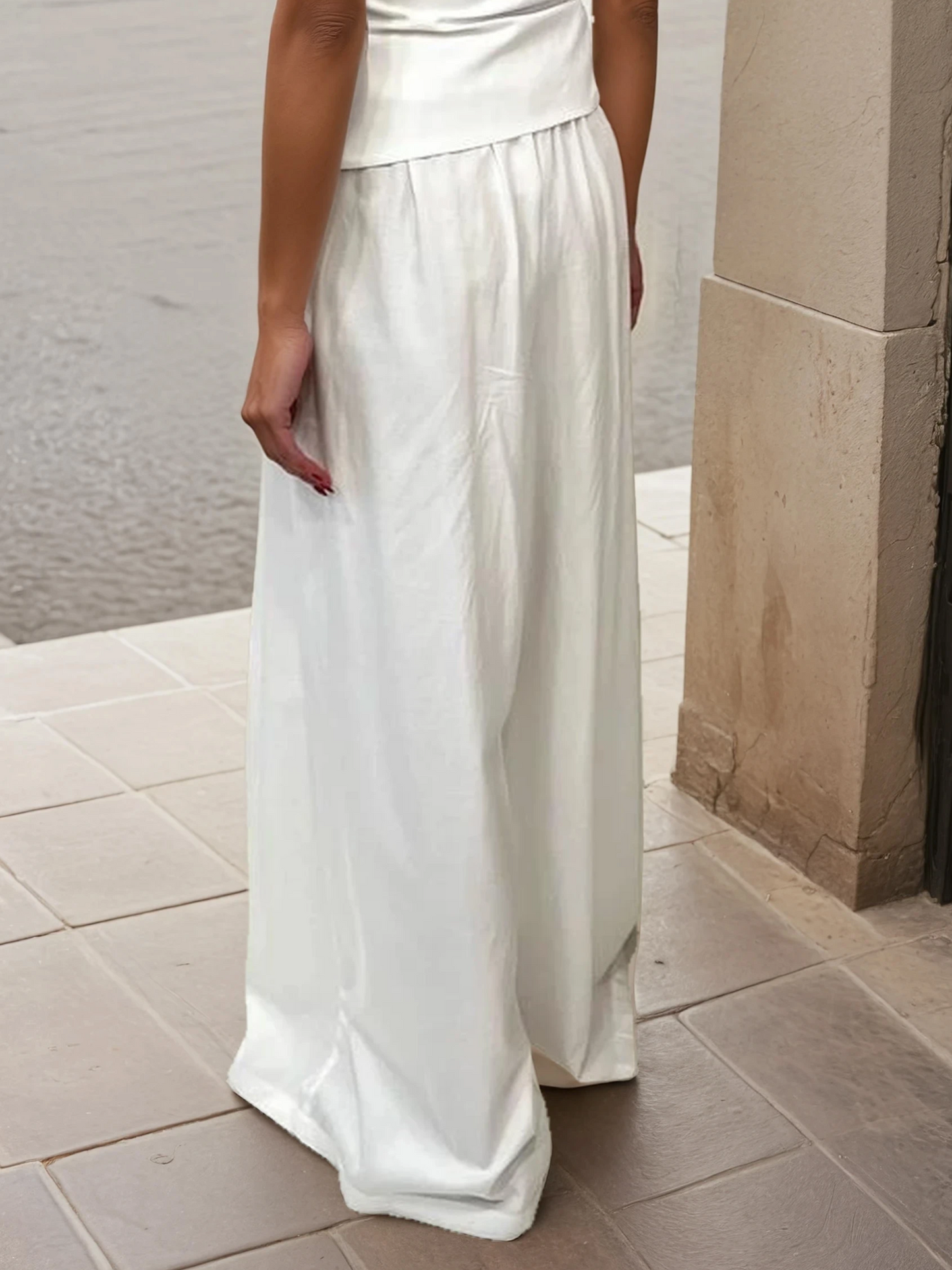 Model wearing ivory wide-leg high-waisted trousers made from a lyocell and cotton blend, styled in an elegant outdoor Mediterranean setting.