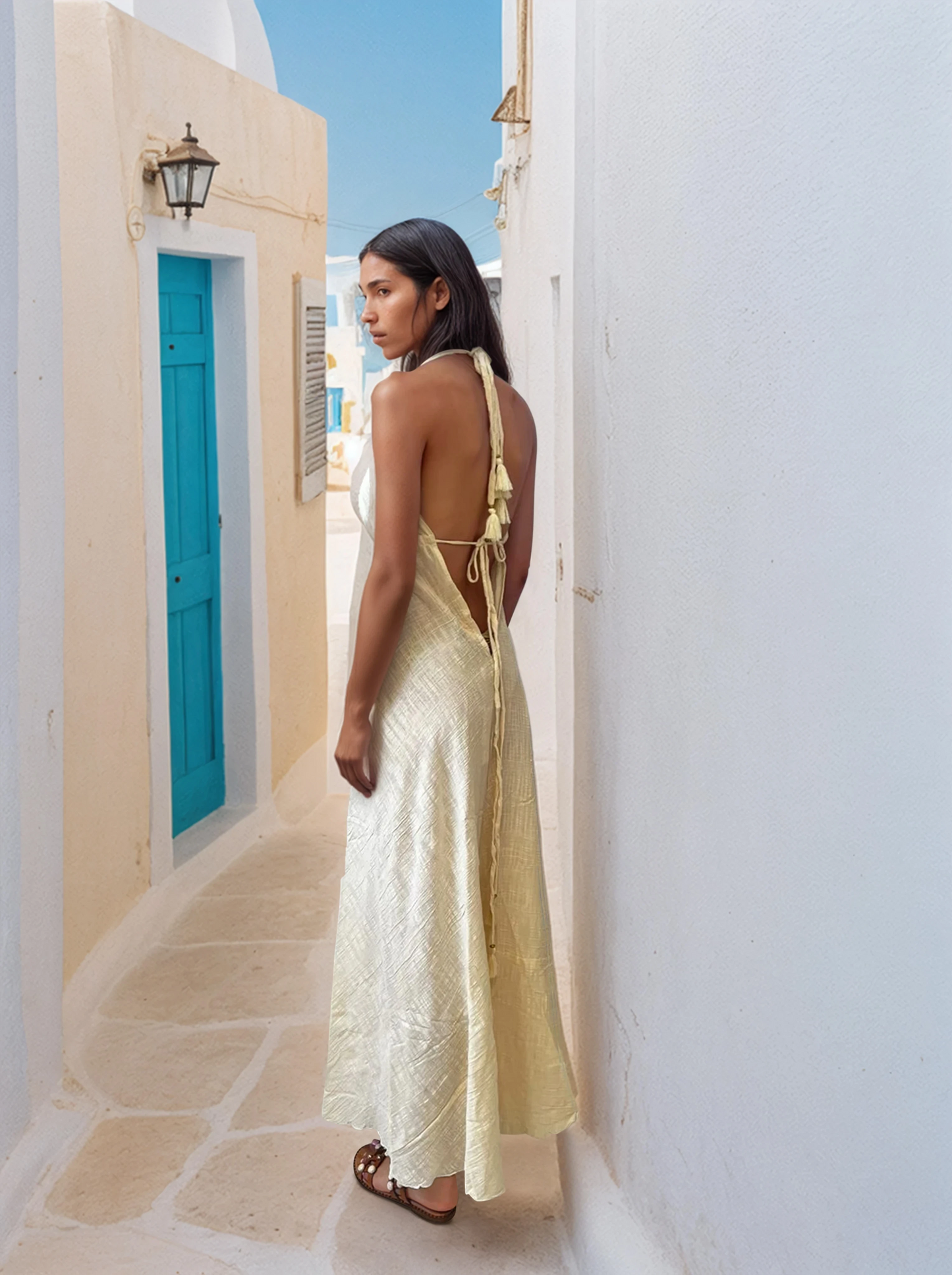 Model wearing a light cotton halter maxi dress with deep neckline and flowing hem, standing on a rooftop terrace with sea view.