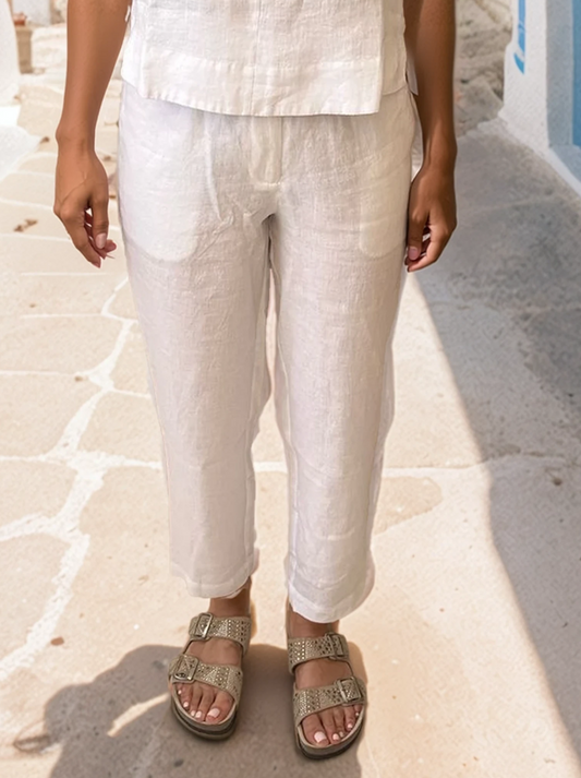 Model wearing relaxed-fit tapered linen trousers with side and back pockets, crafted in Greece from 100% linen, styled in a whitewashed Cycladic alley.